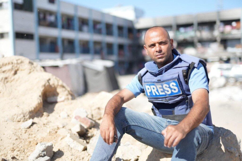 A person wearing a blue vest labeled "PRESS" sitting on rubble in a damaged area. The background includes a partially damaged or abandoned multi-story building, indicating a conflict or disaster zone.